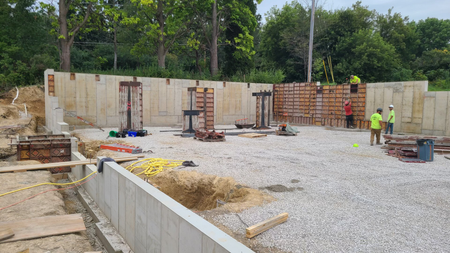 Construction site: Workers building concrete foundation walls. Gravel ground, trees in background.