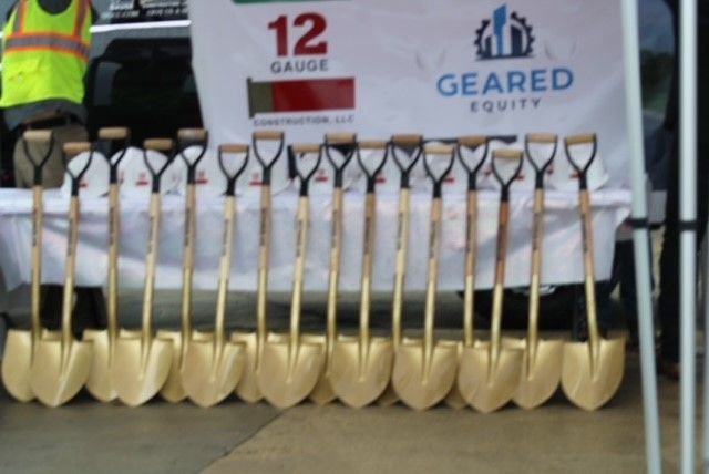 Golden shovels lined up in front of a banner for a groundbreaking ceremony.