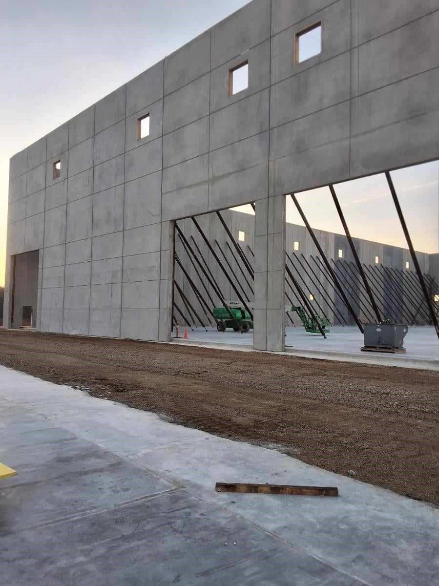 Exterior view of a large concrete building under construction with openings, dirt ground, and a green construction vehicle.