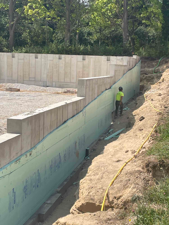 Construction worker on a foundation wall applying insulation. Earth and concrete in outdoor setting.