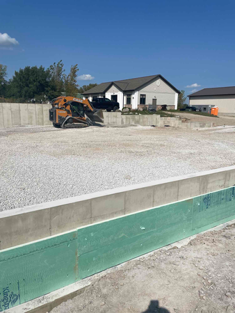 Construction site with a concrete foundation, a skid steer, and a house in the background.