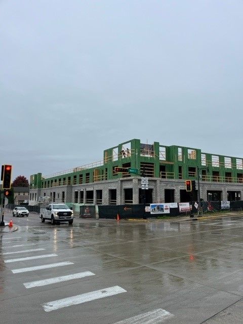 Construction site of a multi-story building on a city street. Grey sky, wet pavement, vehicles present.