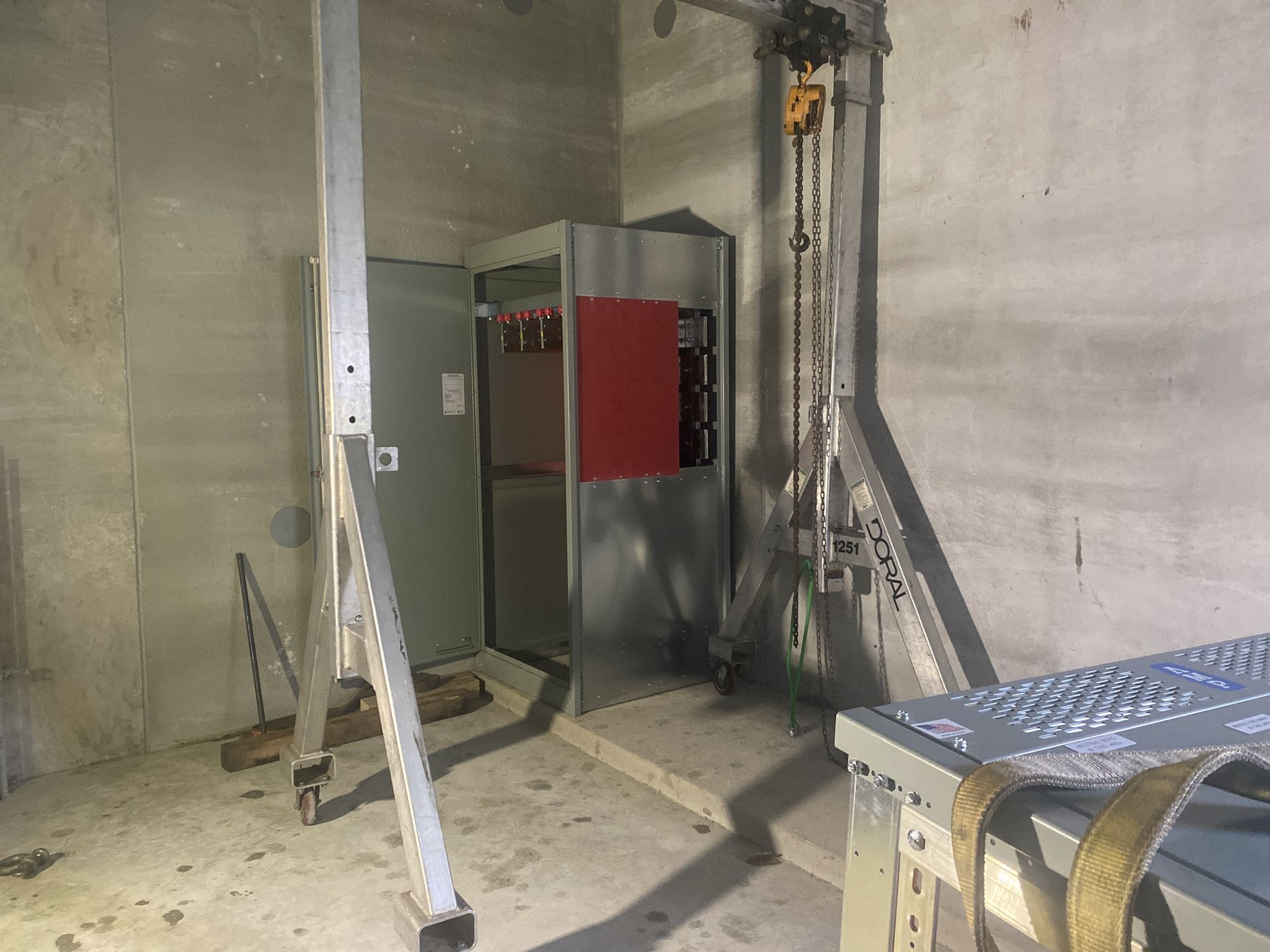 Metal electrical panel, door open, inside a concrete-walled room, supported by metal scaffolding.