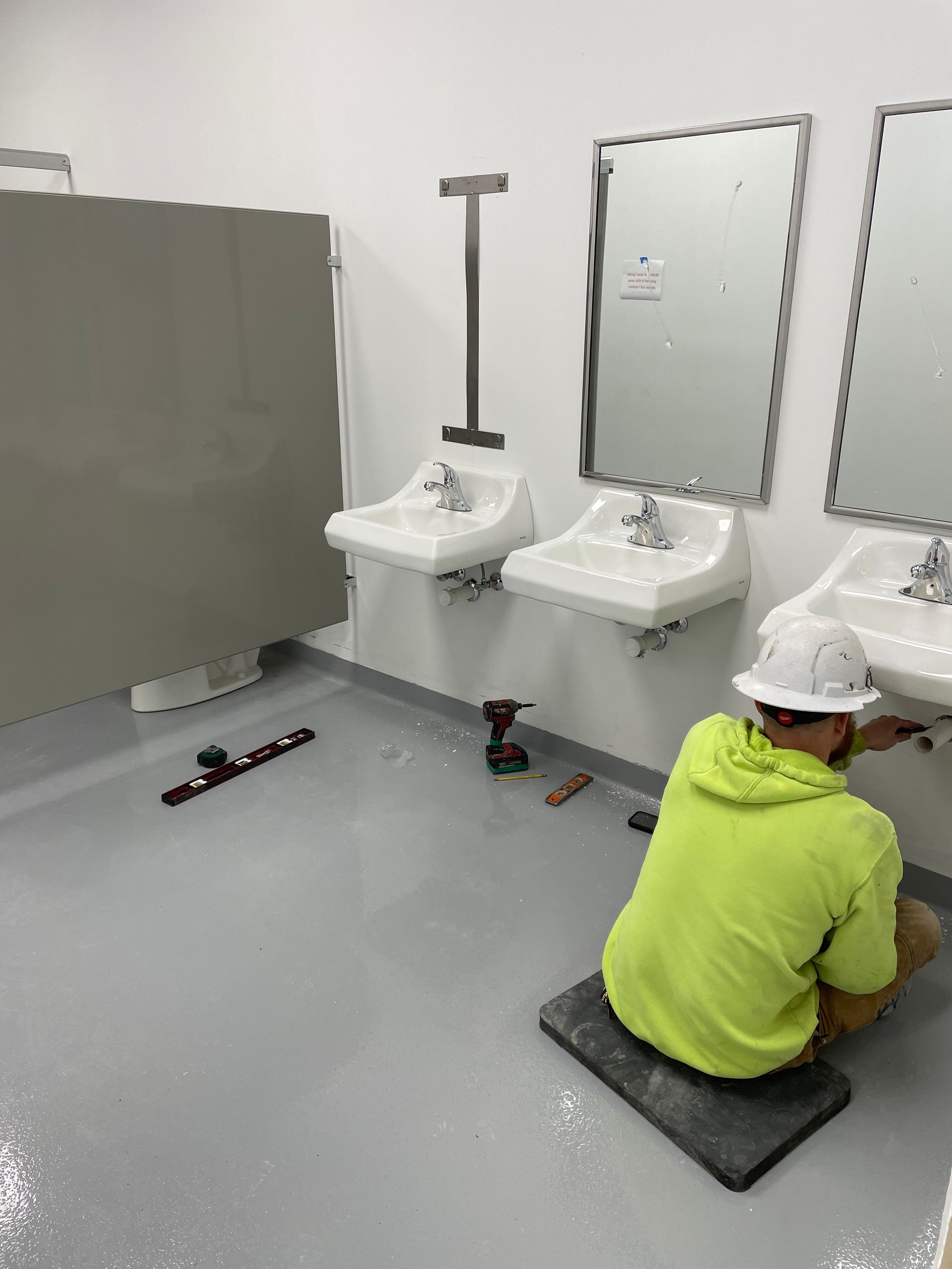 Bathroom with white sinks and stalls; a person in a neon hoodie kneels on the floor cleaning.