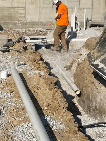 Construction worker in orange shirt and hard hat working on underground pipes in a trench.
