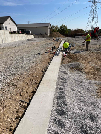 Construction workers installing a concrete curb on a gravel surface next to a building and dirt area.