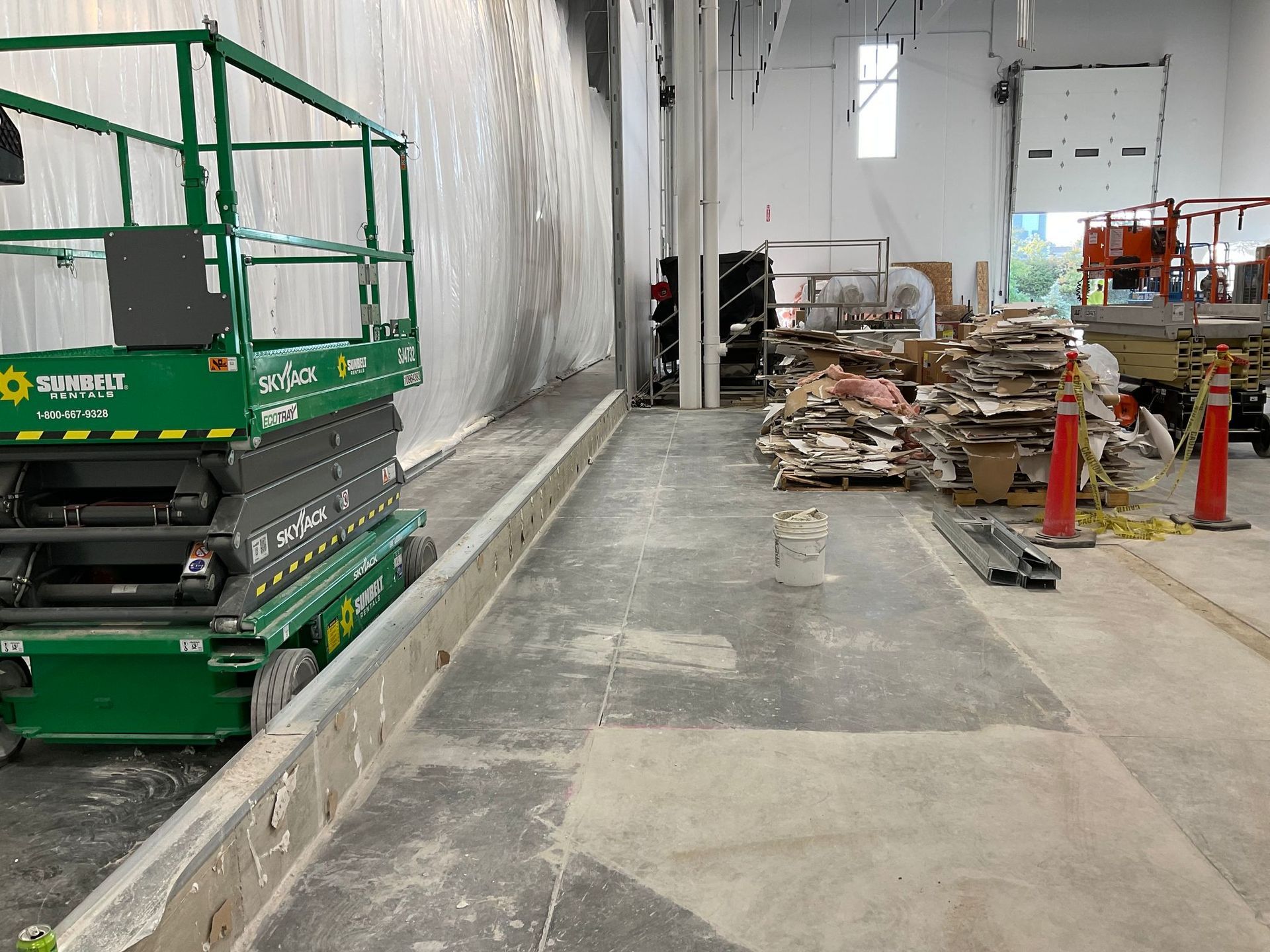 Green scissor lift in an unfinished warehouse. Debris and construction materials are scattered.