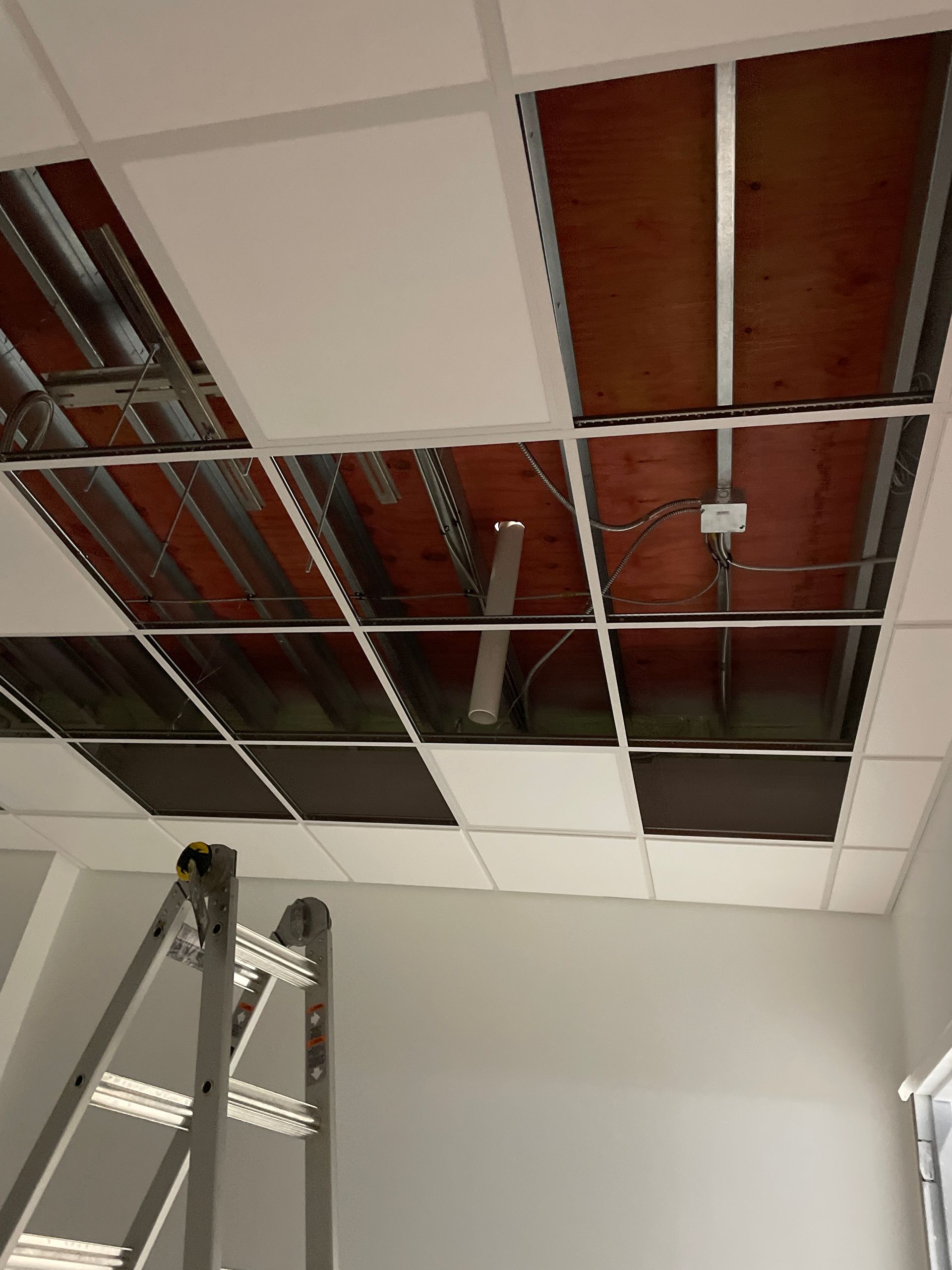 Ceiling with missing drop tiles exposing pipes and wiring above a ladder in a room