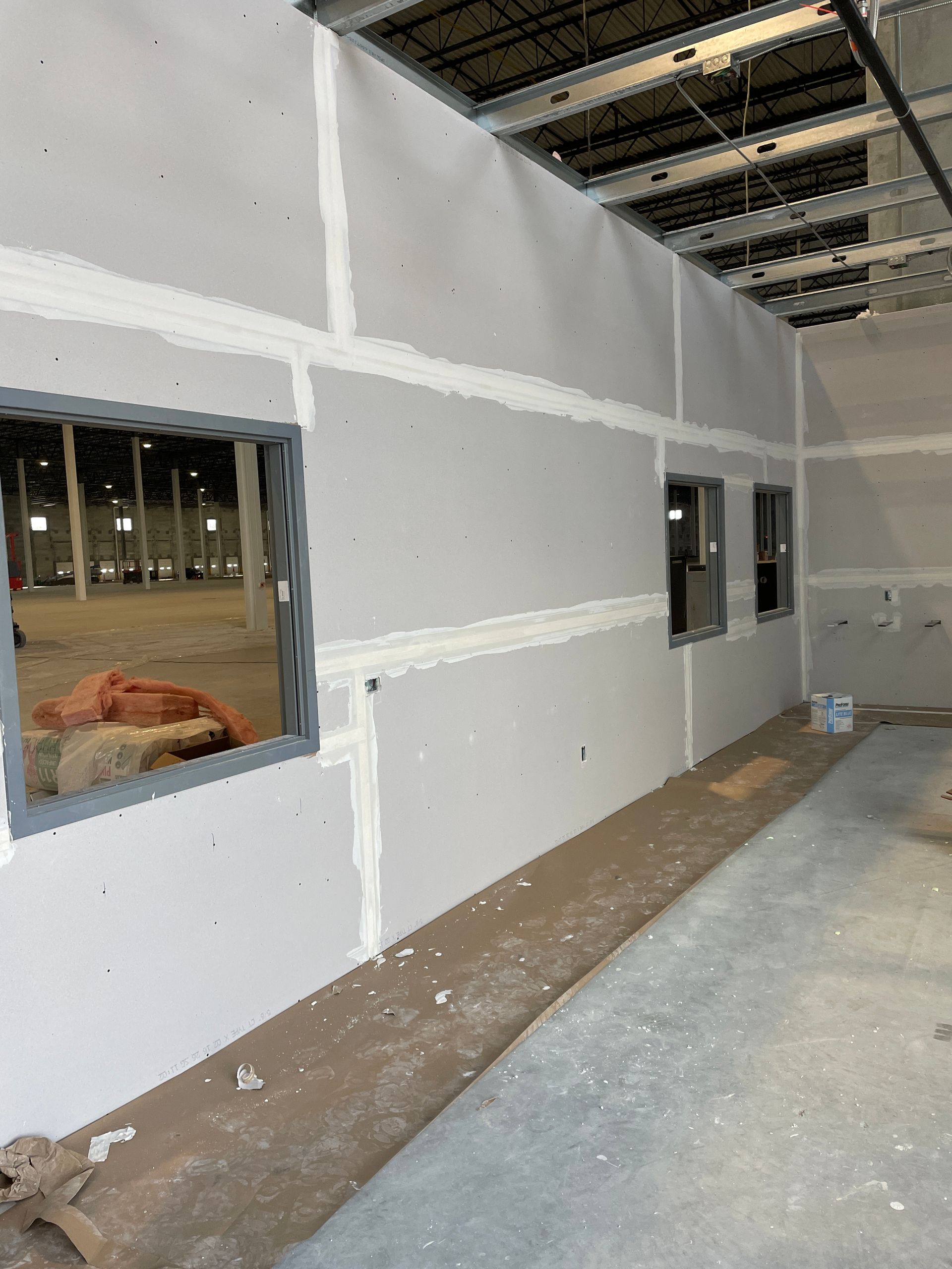 Interior of a building under construction with unfinished drywall, window openings, and exposed ceiling framing.