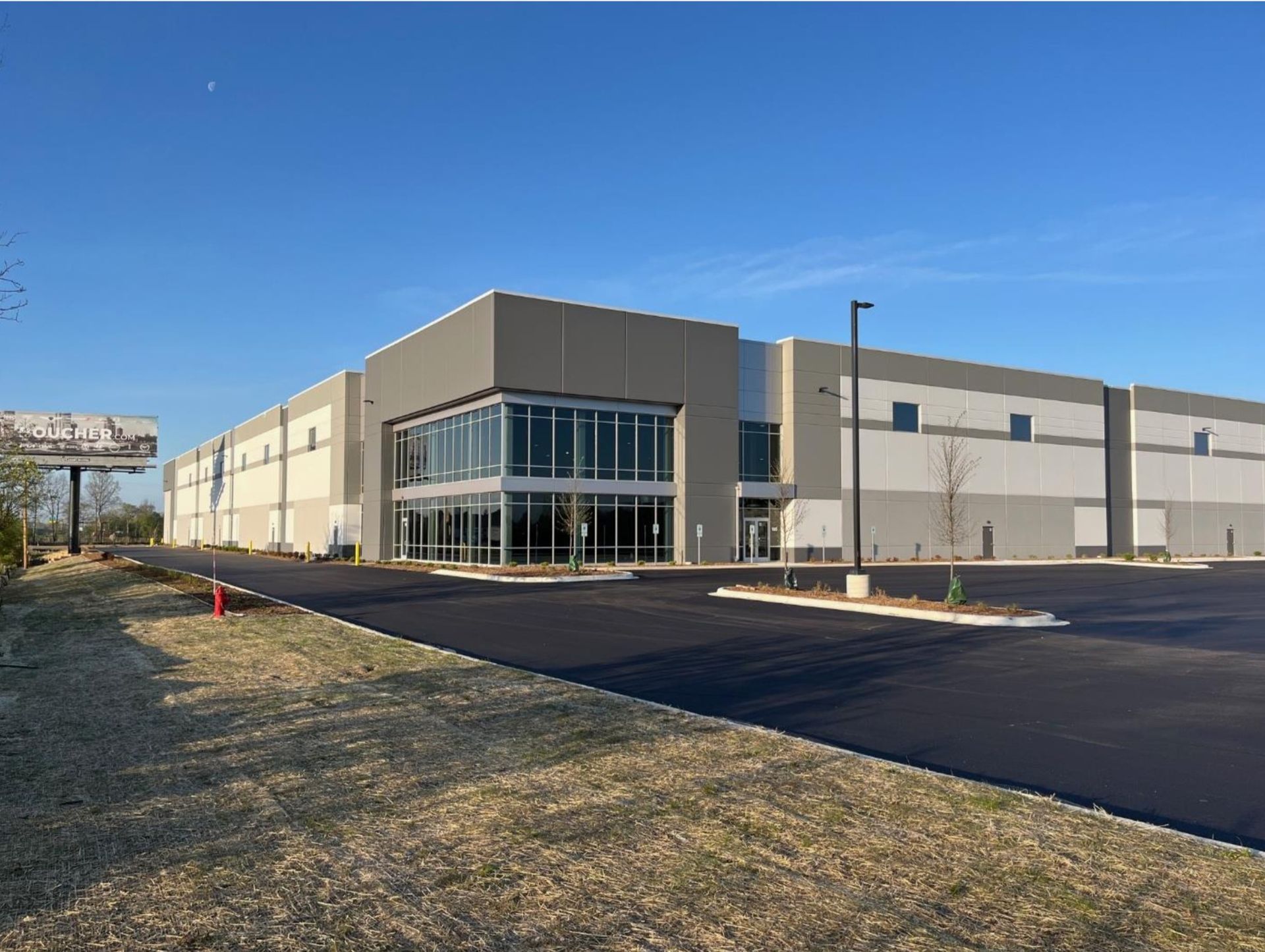 Large beige industrial building with glass corner entrance and new blacktop parking lot under a blue sky