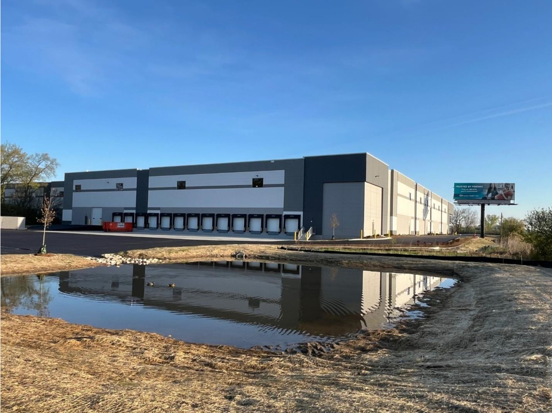 Large white-and-gray warehouse beside a puddle reflecting the building on a sunny day
