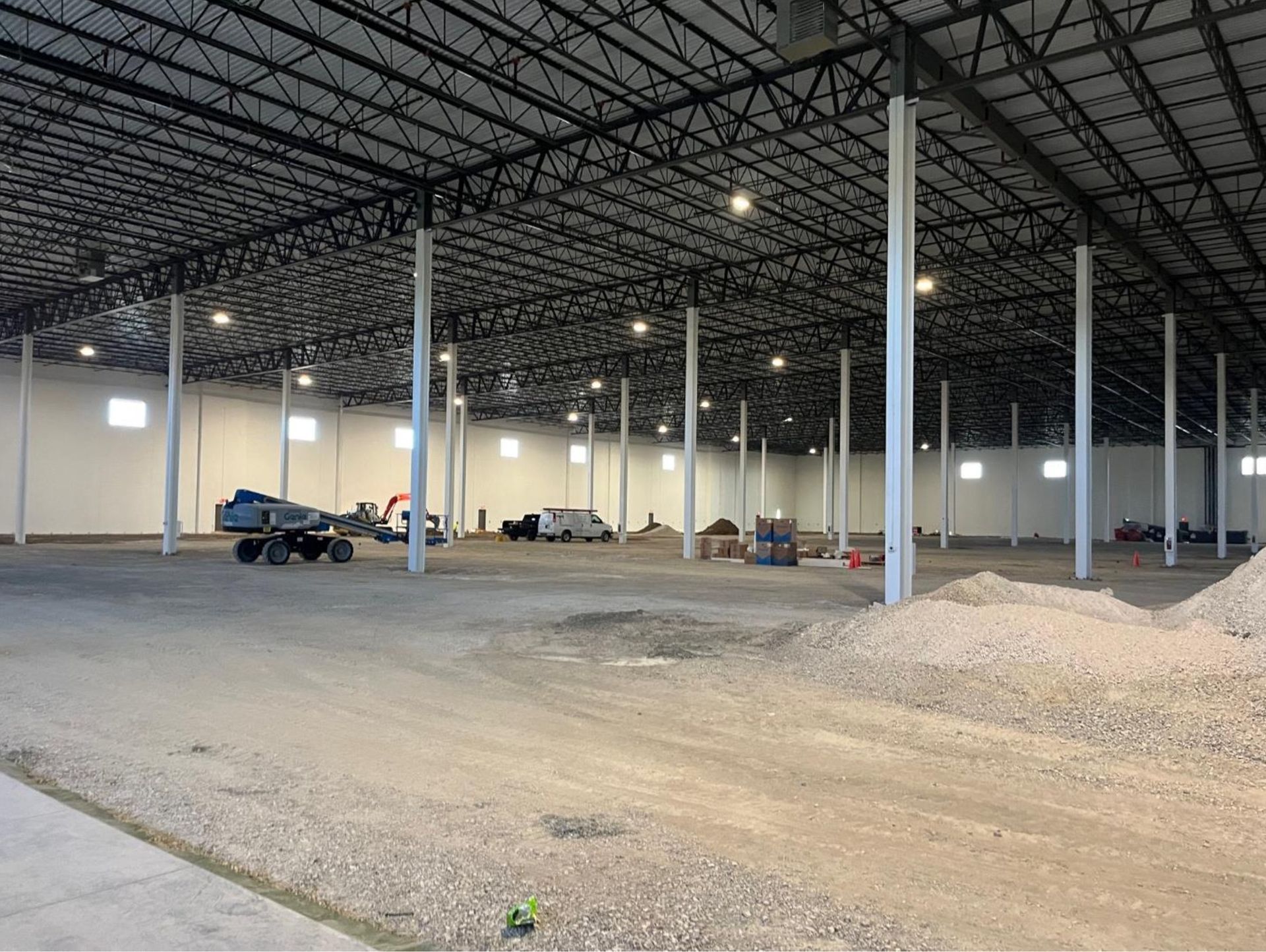Large indoor warehouse under construction with steel roof beams, concrete columns, dirt floor, and a few vehicles inside