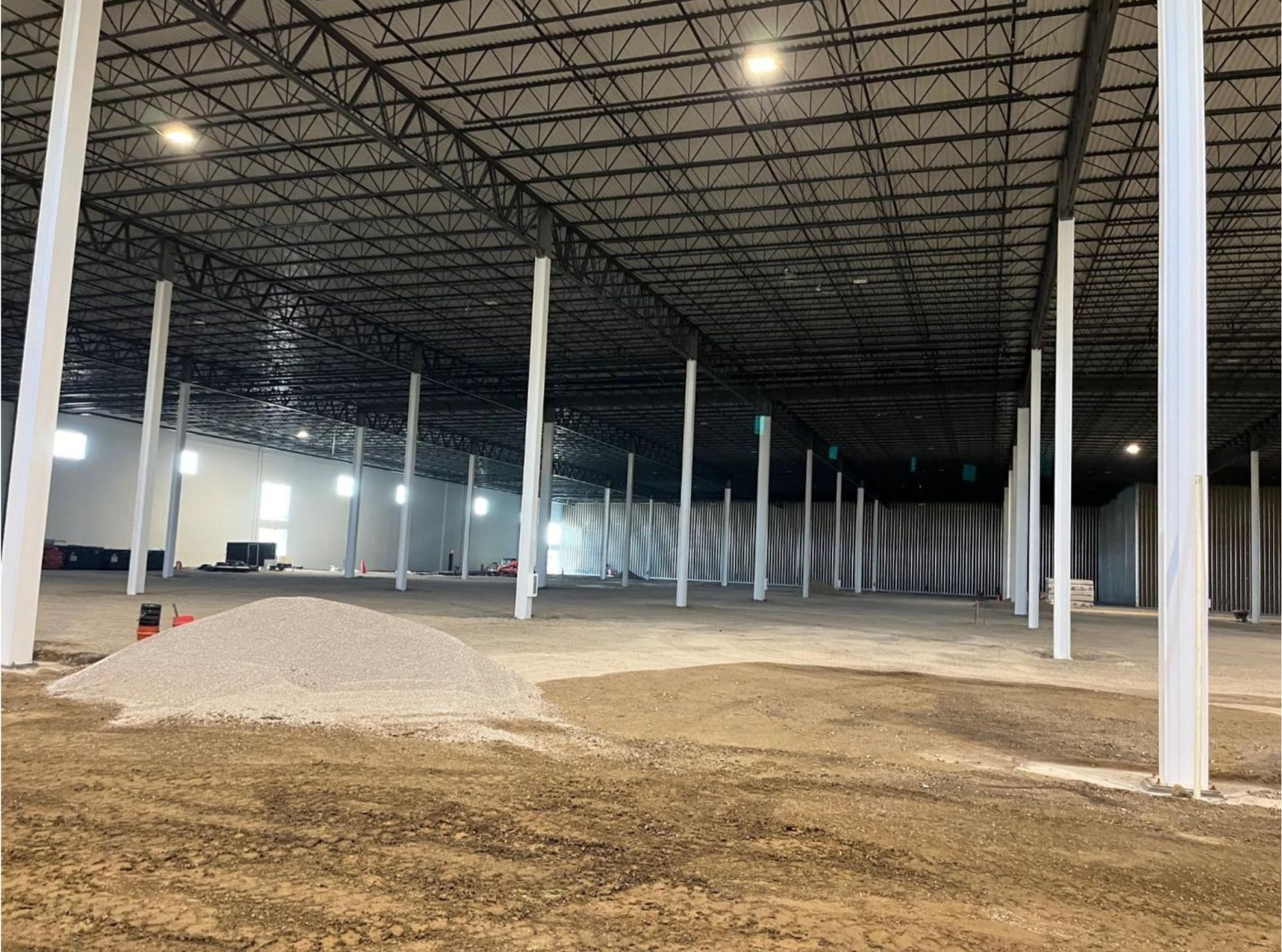 Interior of a large warehouse under construction with rows of white support columns and dirt piles on the floor