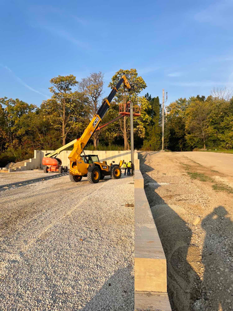Yellow telehandler operating, near a concrete wall and gravel, with a person in the bucket working on a pole.