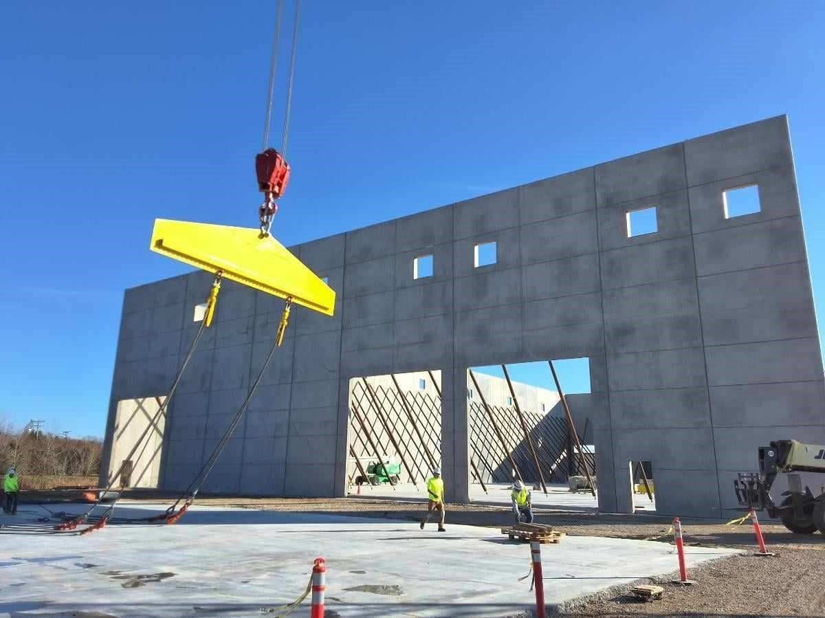 A crane lifting a large yellow concrete panel for a building under construction. Workers are present.