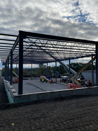 Steel framework of a building under construction, dark blue beams against cloudy sky. Construction workers on site.