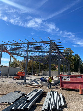 Construction site with steel frame. A worker is near a red dumpster, orange lift, and blue sky.