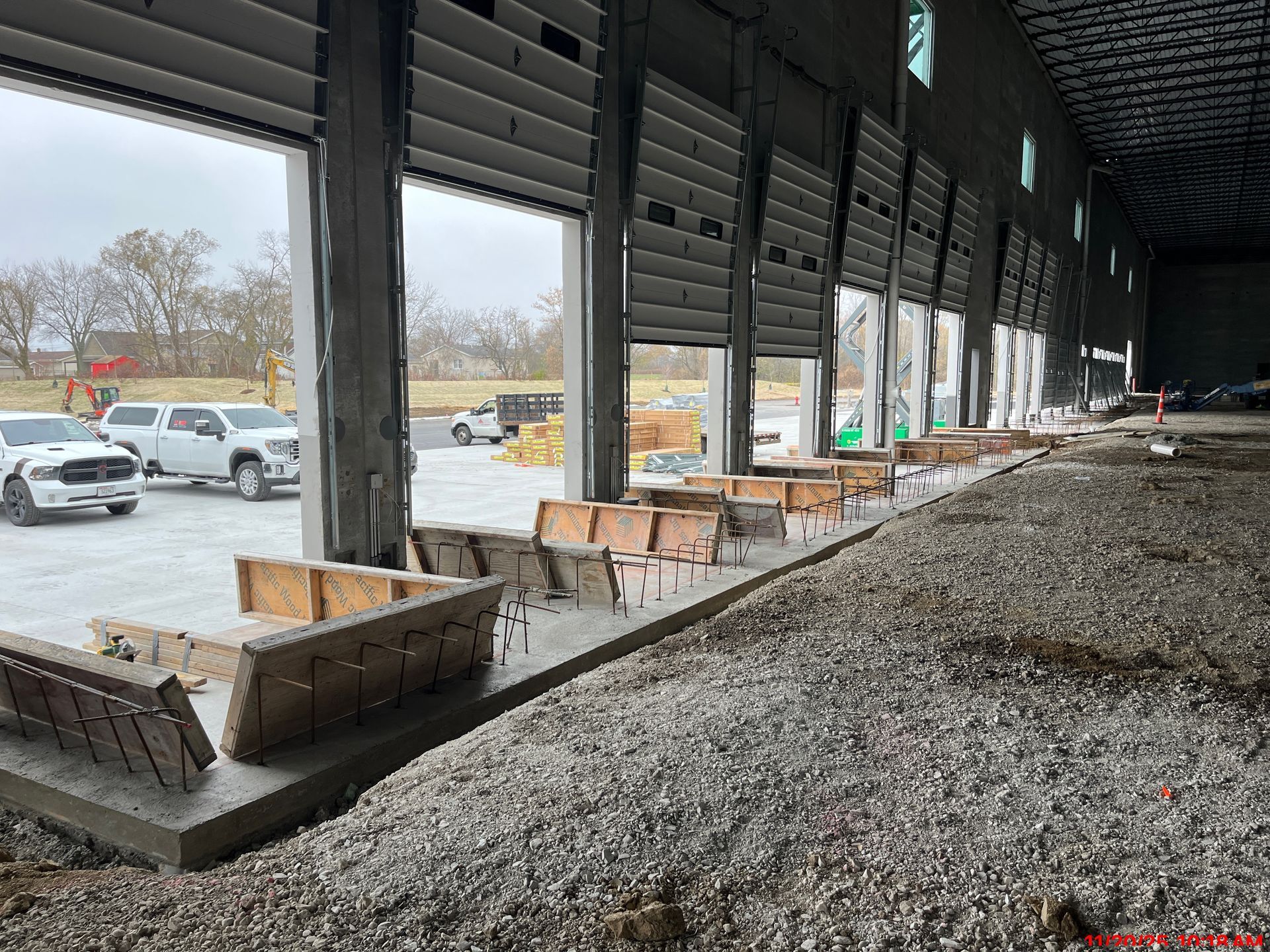 Construction site interior with open loading dock bays, concrete forms, and gravel. Trucks parked outside.