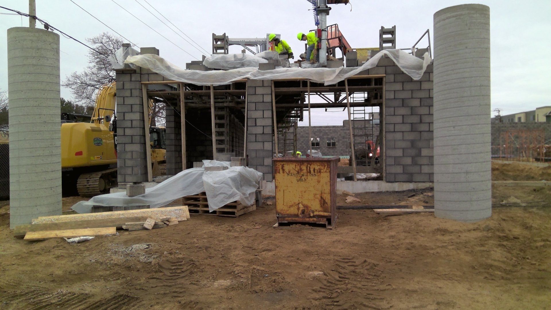 Construction site with workers on a building roof, concrete block walls, and cylindrical concrete pillars.