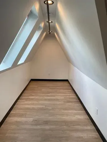 Narrow attic room with angled ceiling, skylights, and wood-look floor. White walls, dark baseboards, and track lighting.
