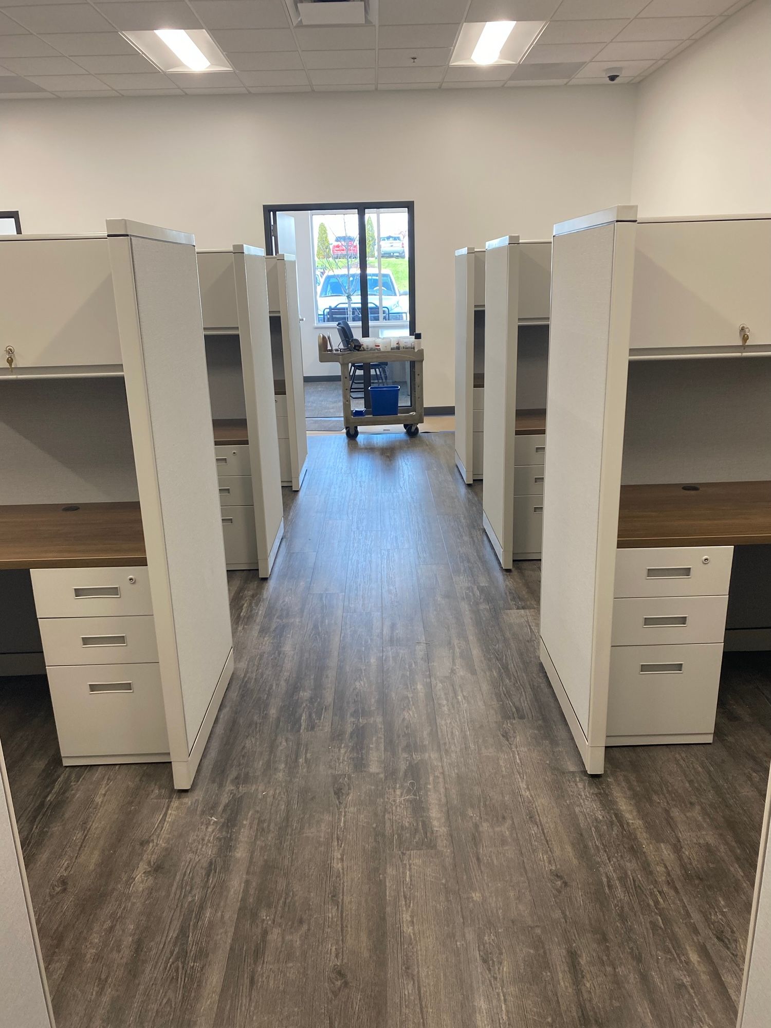 Rows of white office cubicles with wood-toned desktops, set in a brightly lit office.