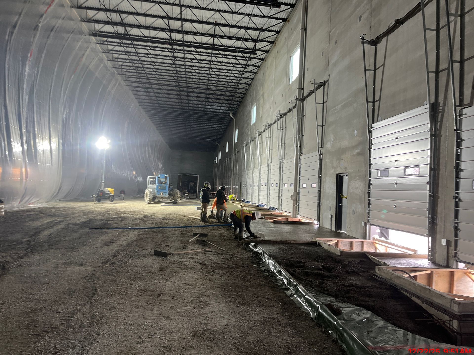 Construction site interior: workers near concrete forms and a long, dark tunnel. Grey walls, exposed metal ceiling.