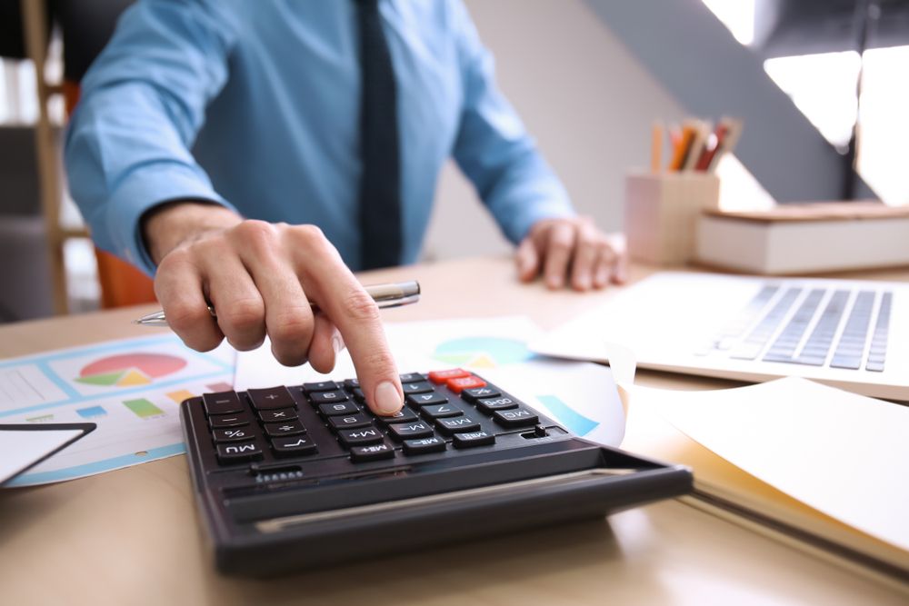 Person in blue shirt using a calculator on a desk with charts and a laptop.