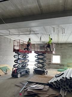 Two workers on scissor lifts, installing pipes on a construction site.