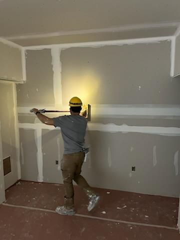 Construction worker taping drywall in a room. He wears a yellow hard hat and is using a taping knife.