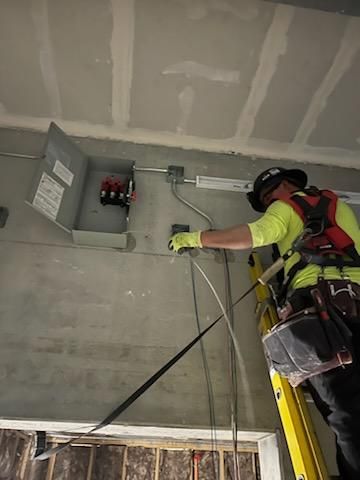 Electrician on a ladder installing wiring near an electrical panel on a construction site.