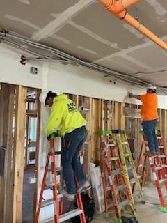 Construction workers installing electrical conduits in a building, using ladders. Orange and yellow shirts.