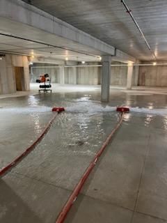 Flooded parking garage with two red hoses spraying water; a pump in the distance.