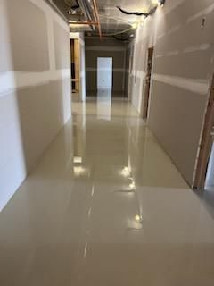 A newly constructed hallway with a reflective, light-colored floor; unfinished walls; and an open doorway at the end.