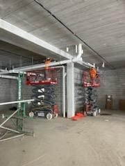 Two workers on scissor lifts install white pipes in a concrete room.