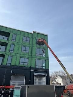 Construction worker in lift basket near building, green siding, gray sky.