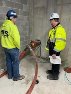 Two workers in safety vests inspect a pipe, likely a water pump, near a wall.