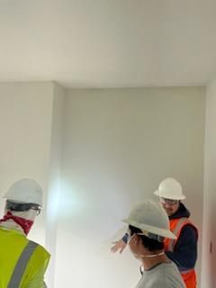 Construction workers in hard hats inspect a room, one points toward the wall.