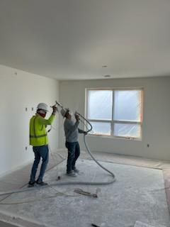 Two construction workers spraying a white substance in a room. One holds the hose, the other operates the sprayer.