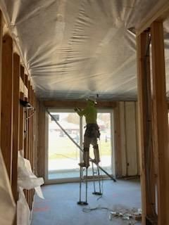 Construction worker on stilts, working on the ceiling of a room under construction, with framing, insulation, and a large window.