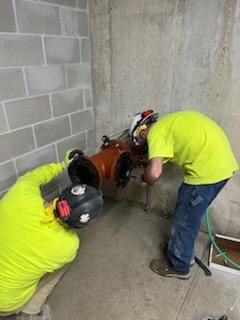 Two workers in neon shirts drill concrete wall. One holds the drill, other assists.