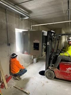 Workers using a forklift to install HVAC unit. One worker guiding; other operating forklift. Concrete wall, construction site.