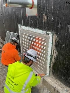 Two workers install metal air vent into a dark wall, applying sealant. One wears orange, the other yellow.
