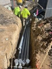 Construction workers in a trench installing grey pipes. Yellow safety vests are worn.