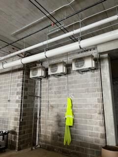 Brick wall with three mounted air conditioning units below pipes and electrical conduit. A yellow safety vest hangs on the wall.