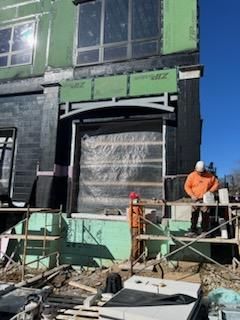 Construction workers on scaffolding, exterior of building under construction. Workers in orange and white attire. Green and black building materials.
