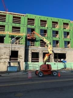 Construction site; orange lift platform near building, workers in it. Bright green sheathing on upper levels. Blue sky.