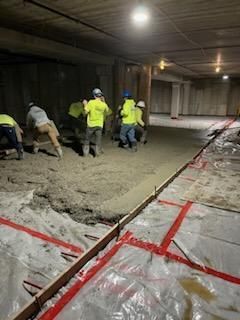 Construction workers pouring concrete on a plastic-covered floor under a concrete ceiling. Yellow vests, white hard hats.