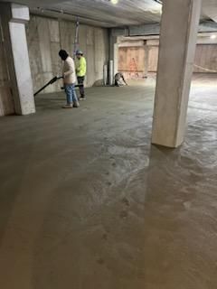 Two workers smoothing a newly poured concrete floor in a large, unfinished room.