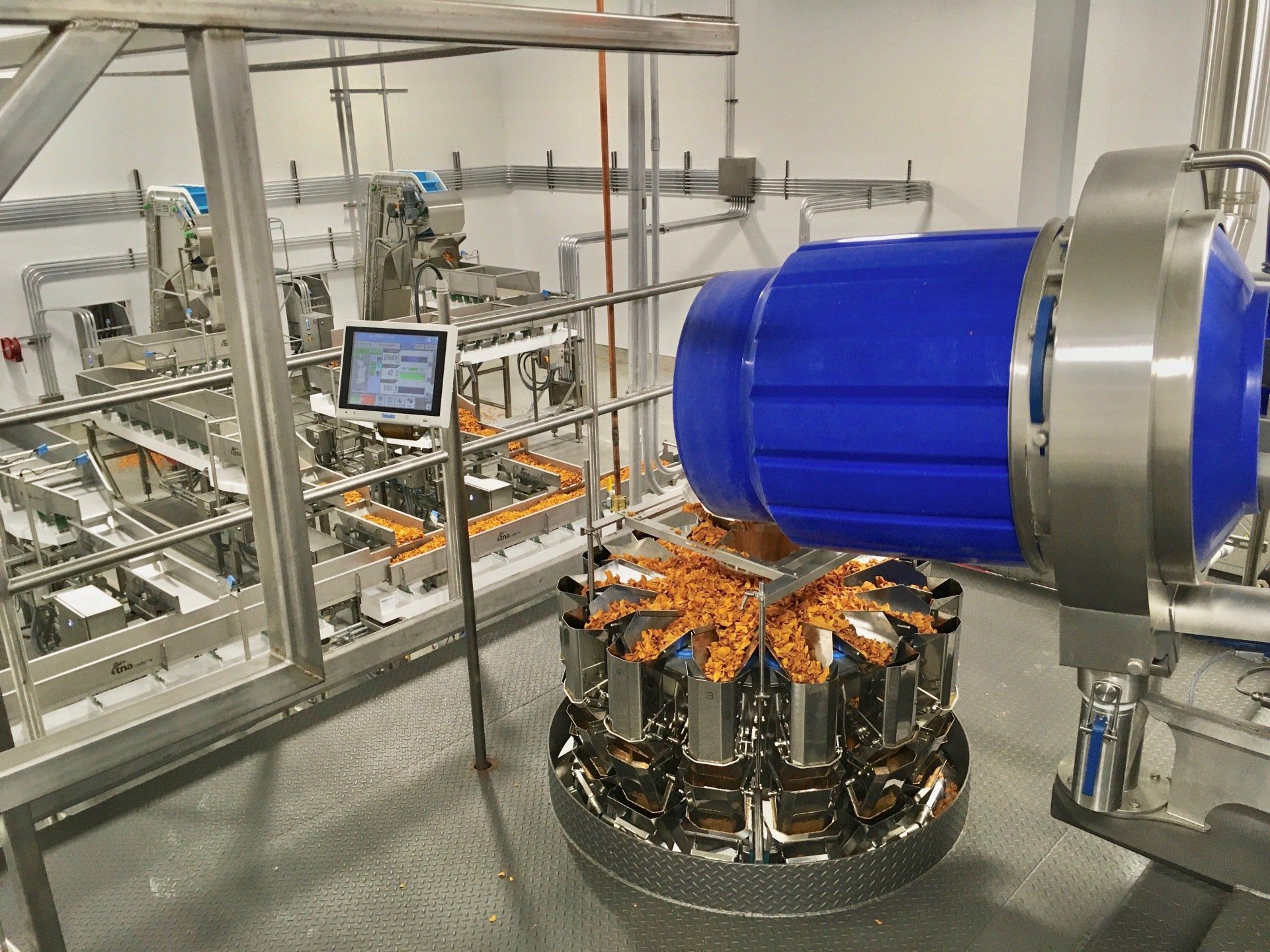Food packaging machine with blue component, conveyor belt, and control screen in a factory setting.