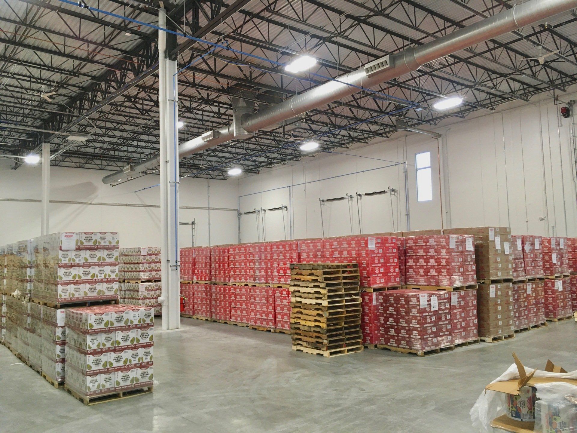 Warehouse interior, stacks of packaged goods on pallets. Red and brown packaging, overhead lighting.